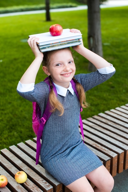 Linda colegial sorrindo, segurando livros e uma maçã na cabeça. feliz em voltar para a escola. rabo de cavalo ruivo.