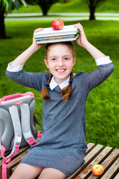 Linda colegial sorrindo, segurando livros e uma maçã na cabeça. feliz em voltar para a escola. rabo de cavalo ruivo.