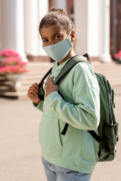 Linda colegiala afroamericana con mochila con máscara protectora