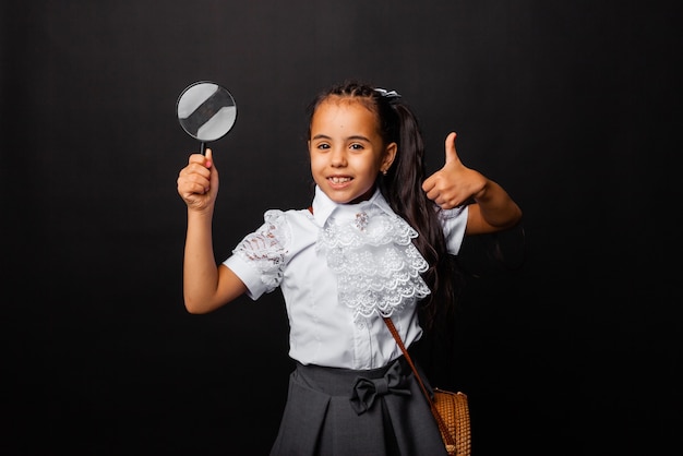 Foto una linda colegiala con cabello largo oscuro sostiene una lupa y muestra la clase sobre un fondo oscuro.