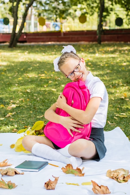 Linda colegiala con gafas sonríe y abraza una mochila mientras está sentada sobre una manta en un soleado parque otoñal. Concepto de regreso a la escuela. Vista vertical