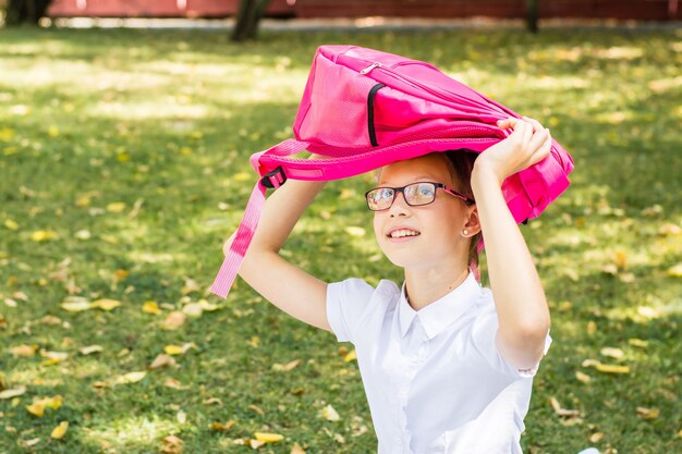 Una linda colegiala con gafas sonríe y sostiene una mochila sobre su cabeza en un soleado parque otoñal. Concepto de regreso a la escuela. Copia espacio