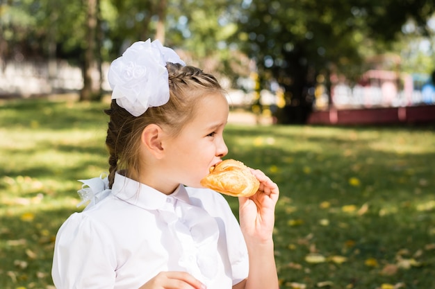 Linda colegiala muerde un trozo de croissant en un picnic en el parque. Comidas del colegio