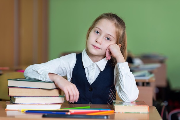 Una linda colegiala se sienta en un escritorio rodeada de libros Mira a la cámara Vida escolar