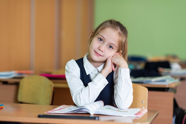 Una linda colegiala se sienta en un escritorio rodeada de libros Mira a la cámara Vida escolar