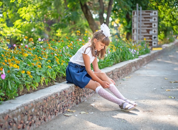 Una linda colegiala con uniforme escolar se sienta y endereza un calcetín en un parque de la ciudad.