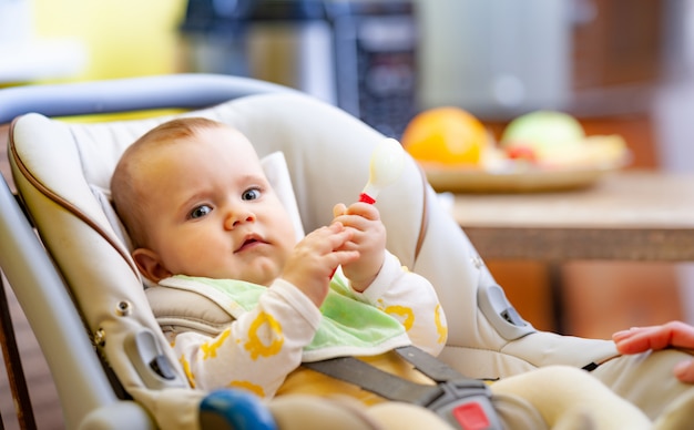 Linda y contenta niña caucásica de seis meses se sienta en un asiento para niños y juega con su amado sonajero.