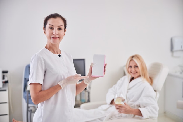 Linda cosmetologista profissional em pé perto do paciente com notebook branco no quarto dentro de casa