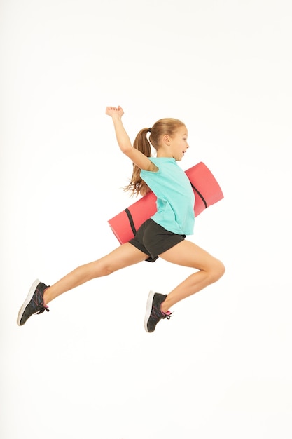 Linda criança do sexo feminino no sportswear segurando rolou o tapete de exercícios e pulando no ar. Isolado em fundo branco