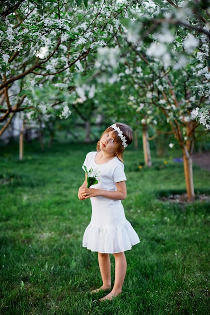 Linda criança garota 5-6 anos segurando flor em pé no jardim de primavera de flor, usando um vestido branco e guirlanda floral ao ar livre, a temporada de primavera está chegando.