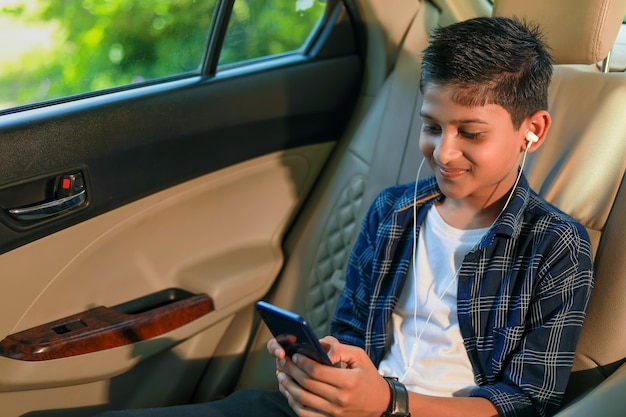 Linda criança indiana sentada no carro usando um smartphone e um gadget de fones de ouvido