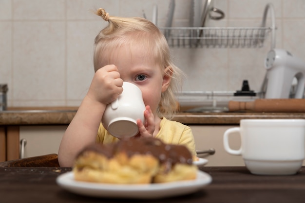 Linda criança loira com um copo nas mãos na cozinha. Menina sorridente bebe um leite. Café da Manhã com Criança