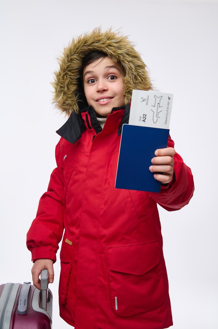 Foto linda criança pré-adolescente, menino branco em uma parca vermelha brilhante com capuz, olhando para a câmera mostrando um passaporte de viagem com cartão de embarque, isolado no fundo branco com espaço de cópia para anúncios