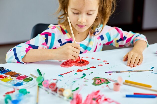 Linda criança sentada à mesa e desenhando um coração vermelho em um papel branco