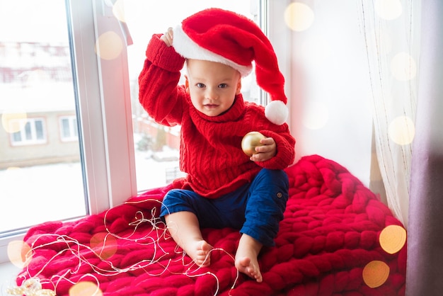 Linda criança sentada perto da janela com chapéu de papai noel vermelho. comemorando o natal em casa