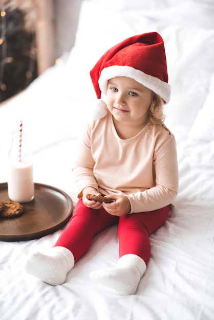 Linda criança sorridente menina 3-4 anos de idade comendo biscoitos de chocolate e bebendo leite fresco em uma garrafa de vidro na cama, sobre o cobertor branco. garoto usa chapéu de papai noel vermelho e pijama em casa. feriados de natal