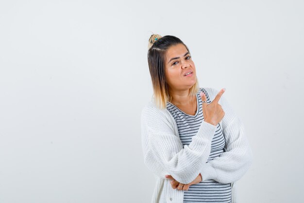 Linda dama en camiseta, chaqueta de punto apuntando a la esquina superior derecha y mirando confiada, vista frontal.