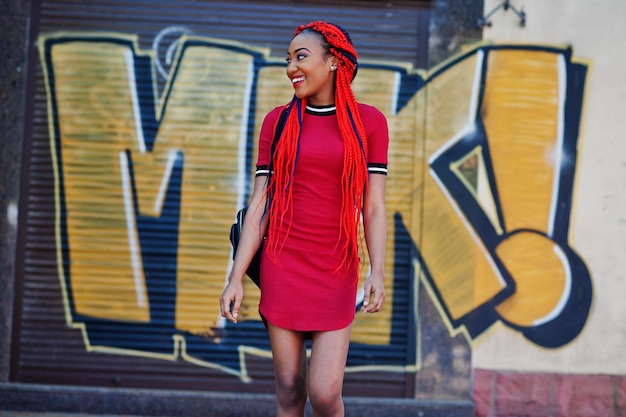 Linda y delgada chica afroamericana en vestido rojo con rastas y mochila posada al aire libre en la pared de graffiti de fondo de la calle Elegante modelo negro