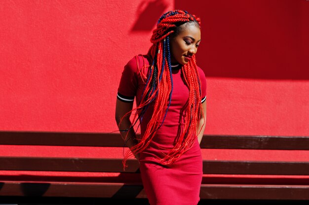 Linda y delgada mujer en vestido rojo con rastas planteadas al aire libre contra la pared roja