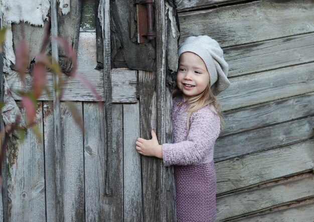 Linda y divertida niña caucásica con abrigo y boina gris mirando por la puerta de madera de la antigua casa de pueblo