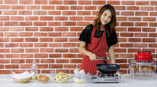 Linda dona de casa asiática com avental listrado vermelho, sorrindo alegremente enquanto cozinha batatas fritas com uma panela preta na mesa da cozinha perto da parede de tijolos que está cheia de utensílios de cozinha, tigelas de vidro e ingredientes