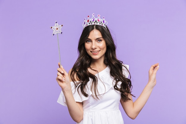 Linda e adorável jovem vestindo roupa de princesa em pé, isolada na parede violeta, posando com uma varinha mágica