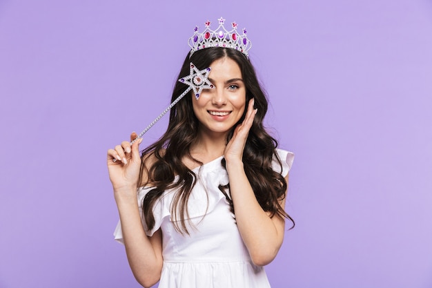 Linda e adorável jovem vestindo roupa de princesa em pé, isolada na parede violeta, posando com uma varinha mágica