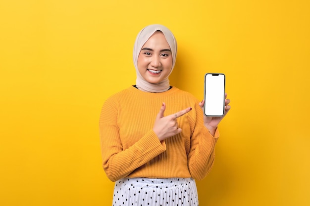 Linda e alegre garota asiática usando hijab apontando o dedo indicador para o celular com tela branca em branco isolada no espaço de cópia de maquete de fundo amarelo