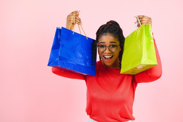 Linda e alegre mulher afro-americana com pilhas de sacos de papel nas mãos se divertindo desfrutando de compras isoladas sobre fundo rosa pastel