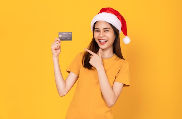 Foto linda e alegre mulher asiática usando chapéu vermelho de natal e segurando cartão de crédito maquete em fundo amarelo