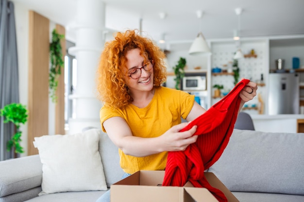 Linda e alegre mulher de cabelo vermelho desembalando um pacote na sala de um novo suéter