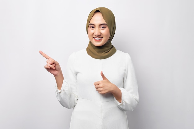 Linda e amigável jovem muçulmana árabe asiática usando hijab apontando o dedo para o espaço vazio da cópia e dando polegares para cima isolado no fundo branco Conceito de estilo de vida religioso de pessoas