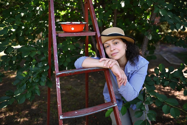 Linda e confiante agricultora caucasiana com chapéu de palha inclinada sobre uma escada em um pomar de cereja e olhando para a câmera