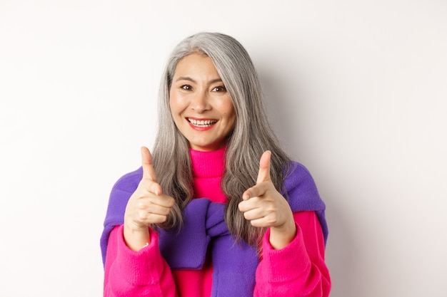 Linda e elegante avó asiática apontando para a câmera, você pode fazer este gesto, parabenizando ou elogiando, em pé sobre um fundo branco