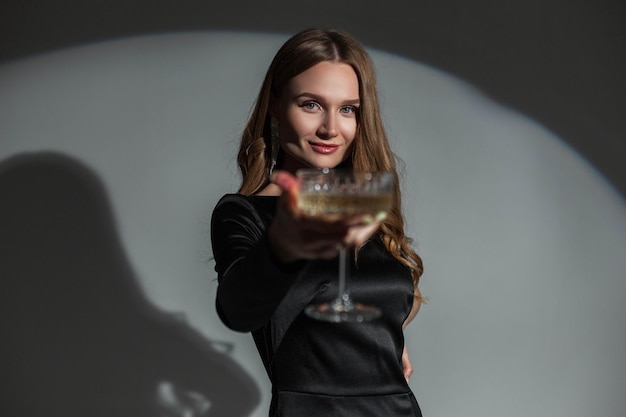 Linda e elegante mulher chique com um sorriso em um elegante vestido preto elegante levanta uma taça de champanhe em um fundo escuro no estúdio Linda senhora na festa