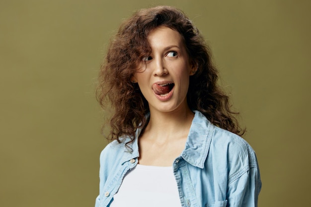 Linda e engraçada fêmea bonita encaracolada em jeans camisa casual enfia a língua para fora posando isolado sobre fundo pastel verde oliva Ser você mesmo Pessoas Estilo de vida emoções conceito Espaço para texto