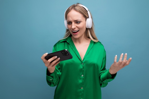 Linda e entusiasmada garota loira atraente com fones de ouvido e smartphone em fundo azul