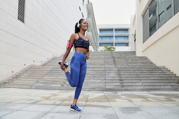 Linda e feliz desportista afro-americana com fones de ouvido, apoiada em uma perna só e alongando os quadríceps