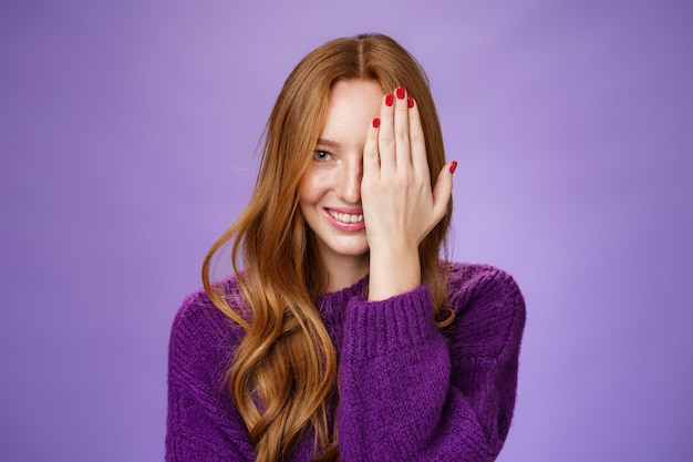 Linda e feliz garota ruiva atraente espiando com um olho cobrindo metade do rosto com a palma da mão, sorrindo sonhadora de felicidade e humor brincalhão posando encantada e despreocupada sobre fundo roxo.