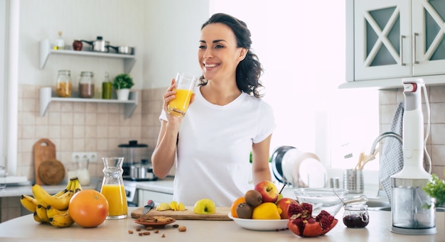 Foto linda e feliz jovem morena na cozinha em casa está bebendo suco de laranja
