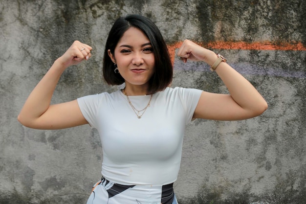 Linda e forte jovem asiática com camisa branca mostrando bíceps e sorridente garota indonésia forte conceito