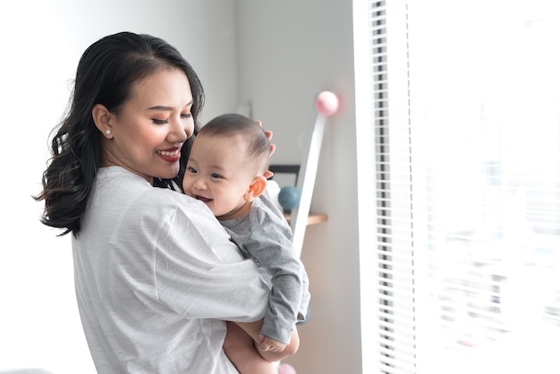 Linda e jovem mãe está brincando com seu lindo bebê e sorrindo enquanto está perto da janela em casa