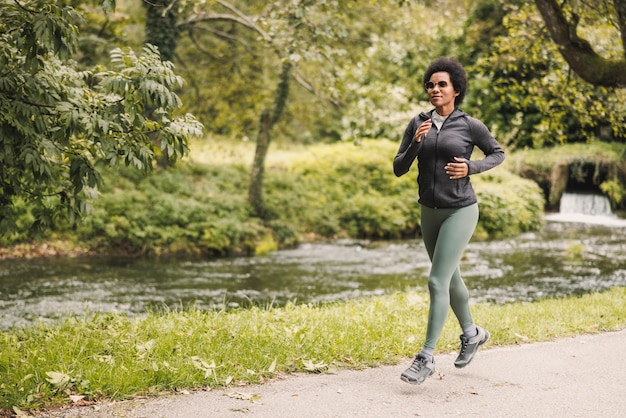 Linda e madura mulher afro-americana sorridente está correndo ao longo da trilha perto do rio das montanhas na natureza.