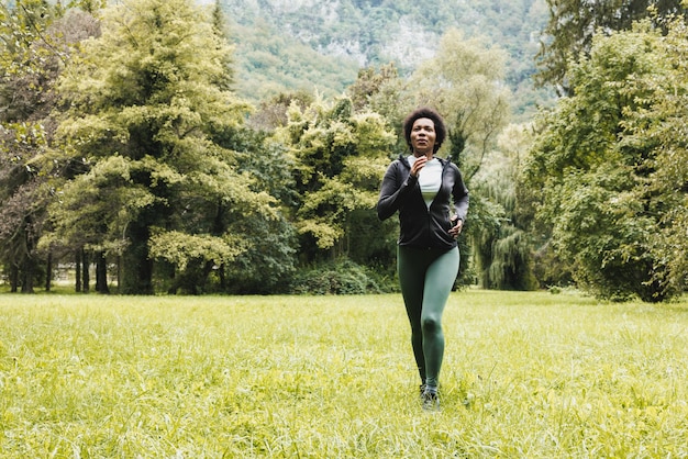 Linda e madura mulher afro-americana sorridente está correndo pela grama verde perto da paisagem montanhosa.