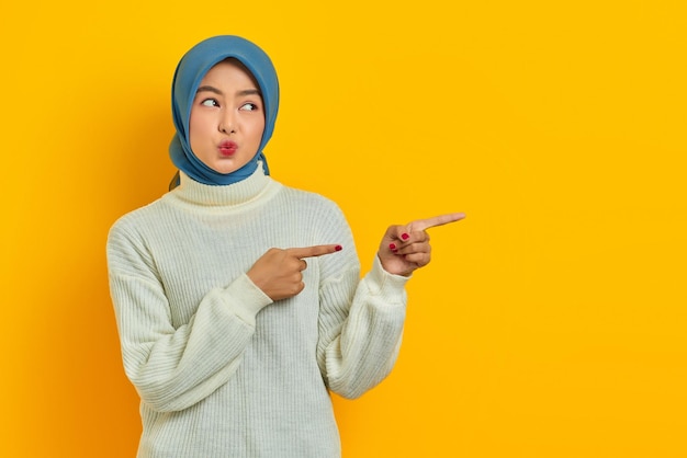 Linda e pensativa mulher asiática de suéter branco parece pensar seriamente em perguntas e apontar dedos isolados sobre fundo amarelo