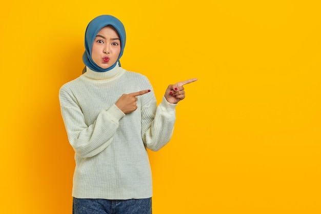 Linda e pensativa mulher asiática de suéter branco parece pensar seriamente em perguntas e apontar dedos isolados sobre fundo amarelo