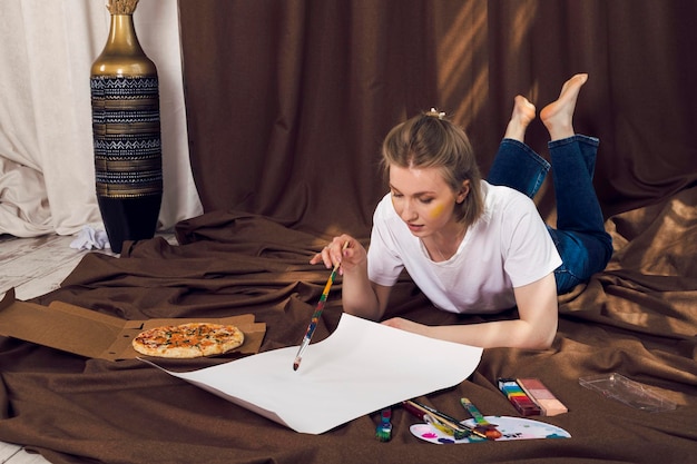 Foto linda e relaxada pintora ou artista loira deitada em um pano marrom na confortável oficina comendo pizza e pintando sob os raios do sol caindo sobre ela