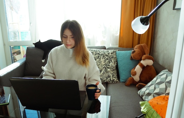 Linda e séria jovem sentada no sofá com brinquedos macios e trabalhando com um laptop em casa com uma xícara de café o conceito de trabalho remoto em casa