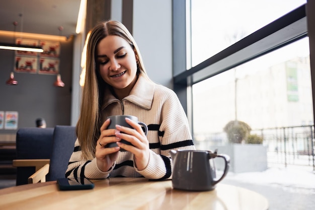 Linda e sorridente jovem calma tomando café olhando pela janela enquanto está sentado em uma mesa em um restaurante Café da manhã