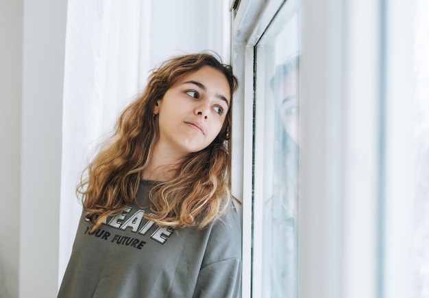 Foto linda e triste adolescente infeliz com cabelo encaracolado, sentado no peitoril da janela